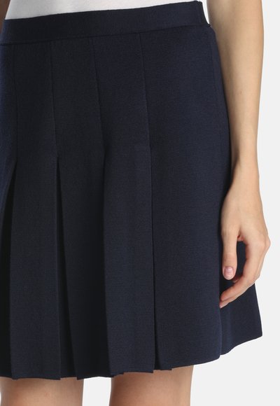 Ręka kobiety spoczywająca obok granatowej plisowanej spódnicy, z włożoną białą bluzką, widoczna od talii do połowy uda na jednolitym tle.