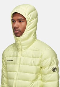 Helle gelbe isolierte Jacke mit gestepptem Design, durchgehendem Reißverschluss und enganliegender Kapuze. Mit schwarzem Logo auf Brust und Ärmel für Branding.