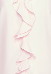 Blusa rosa chiaro con un dettaglio a volant verticale lungo un lato. Il tessuto appare liscio con una consistenza morbida. Nessun ulteriore decoro.
