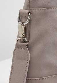 Šedá semišová taška se stříbrným kovovým zapínáním, která má texturovaný povrch a vícestupňové prošívání na látkovém popruhu.