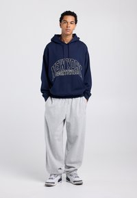 Sweat à capuche bleu marine avec les inscriptions "NEW YORK" et "SPORTSWEAR" en crème, associé à un pantalon de jogging gris clair. Les chaussures comportent des accents bleus et blancs.