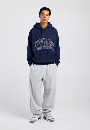 Laivastonsininen huppari, jossa on vaalean kermanväriset "NEW YORK"- ja "SPORTSWEAR"-kuviot, yhdistetty vaaleanharmaisiin juoksuhousuihin. Jalkineissa on sinisiä ja valkoisia yksityiskohtia.