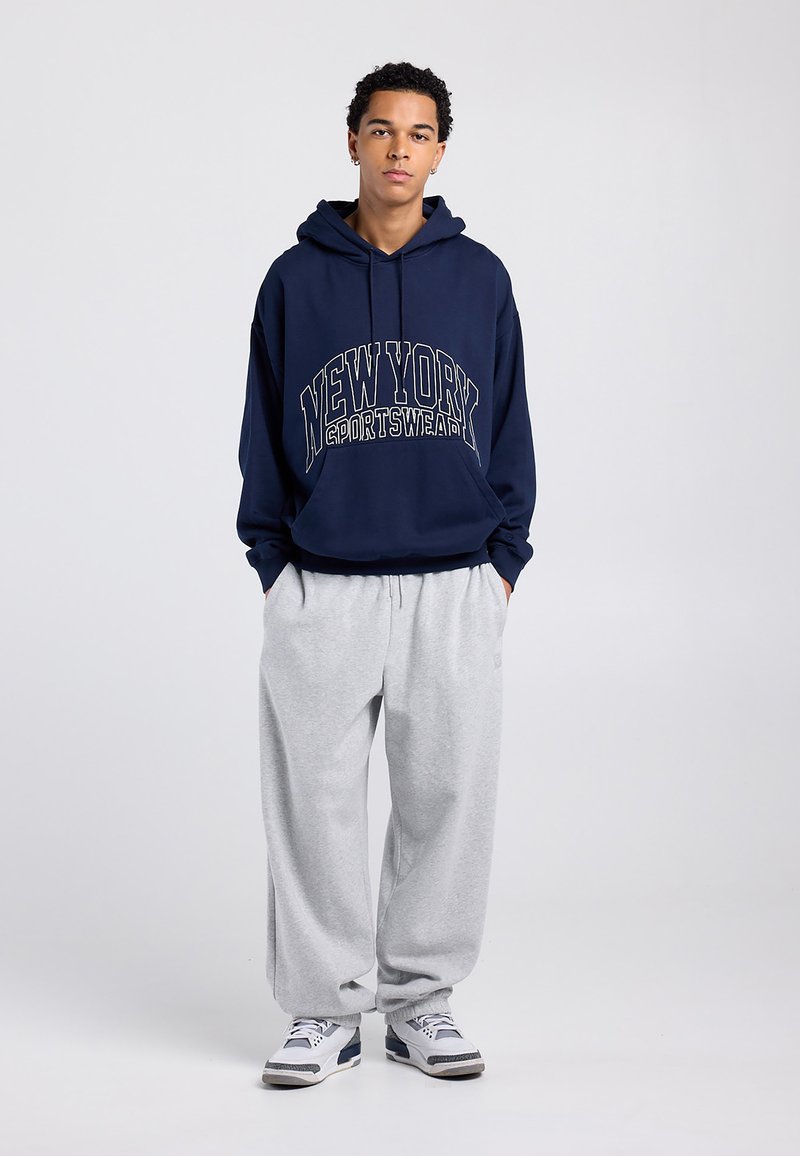 Navyblauer Kapuzenpullover mit den Aufdrucken "NEW YORK" und "SPORTSWEAR" in Creme, kombiniert mit hellgrauen Jogginghosen. Die Schuhe haben blaue und weiße Akzente.