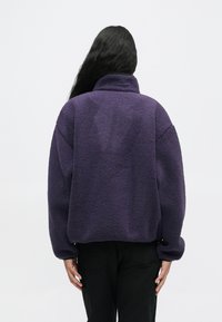 Pikkade mustade juustega inimene, kes kannab lilla fliisjakki ja musti pükse, on seljaga lihtsa heleda tausta poole.