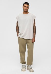 Lichtgrijs mouwloos topje met een ronde hals, gecombineerd met beige joggingbroek en lichtgrijze sneakers. Bevat tatoeages op de armen.