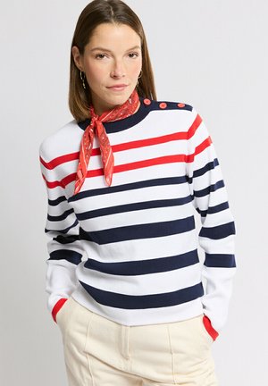 Femme portant un pull blanc avec des rayures rouges et bleu marine, des boutons rouges sur les épaules, un foulard rouge à motifs autour du cou, et un pantalon beige clair, mains dans les poches.