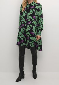 Vestido negro con un vibrante estampado floral en verde y morado, de manga larga y diseño de botones, combinado con leggings de cuero negro y botines.
