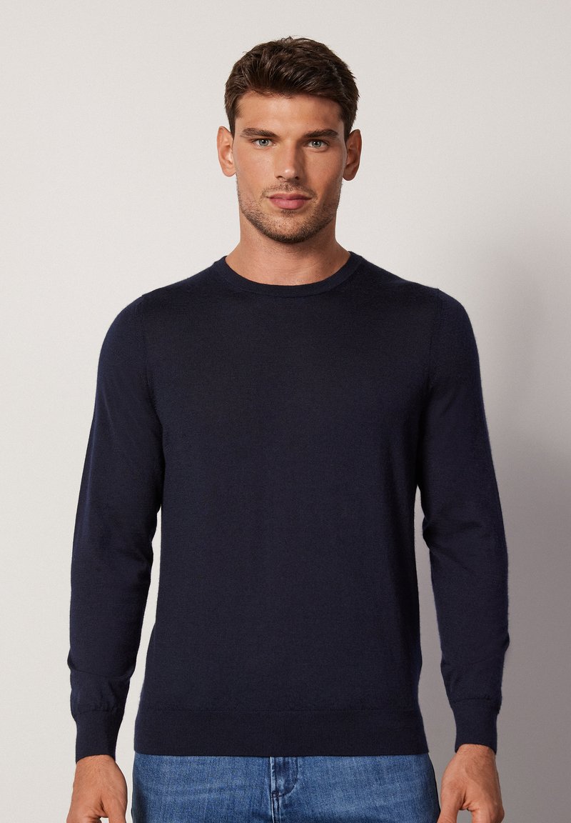 Navy blue crew neck sweater, made of soft fabric, featuring long sleeves and ribbed cuffs, worn with light blue denim jeans.