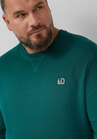 Zielona bluza wykonana z bawełny, z okrągłym dekoltem i ściągaczami na mankietach. Posiada małe kwadratowe logo po lewej stronie. Gładka tekstura.