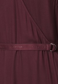 Robe portefeuille en tissu bordeaux avec ceinture assortie ornée du logo "Calvin Klein" embossé et boucle double anneau argentée.