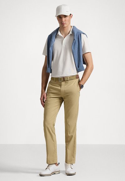 Chinos bege, camisa polo branca, suéter azul colocado sobre os ombros, boné branco e sapatos brancos com detalhes castanhos. Relógio circular no pulso.