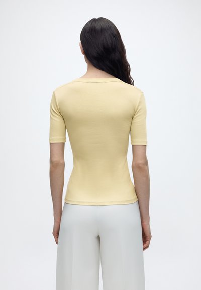 Γυναίκα με μακριά σκούρα μαλλιά που φορά εφαρμοστό ανοιχτό κίτρινο κοντομάνικο τοπ και ανοιχτόχρωμο παντελόνι με φαρδιές μασχάλες, γυρισμένη με την πλάτη σε απλό φόντο.