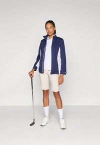 Mornarska golf jakna z belimi stranskimi paneli, bela polo majica, bež kratke hlače, bele nogavice in golf palica, postavljeni na enobarvno ozadje.