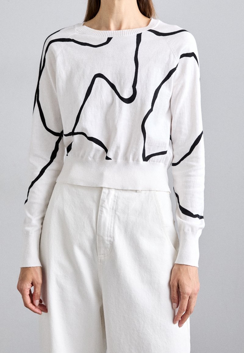 Femme portant un pull à manches longues blanc avec des motifs abstraits en lignes noires et un pantalon blanc taille haute, sur un fond gris uni.