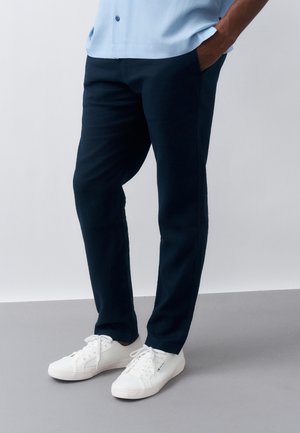 Άνδρας που φοράει σκούρο μπλε παντελόνι, ανοιχτό μπλε πουκάμισο και λευκά αθλητικά παπούτσια, στέκεται με τα χέρια στις τσέπες σε γκρι πάτωμα και λευκό φόντο.