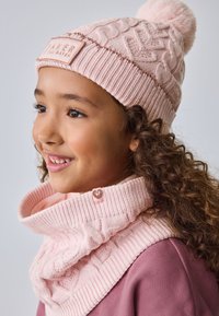 Svetlo roza pletena kapa z vzorcem kaveljčkov in cofkom, skupaj z ujemajočo se pleteno ruto z detajlom gumbka v obliki srca.