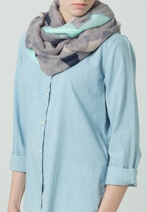 Personne portant une chemise bleu clair à boutons avec les manches retroussées et une écharpe à motifs multicolores enroulée autour du cou.