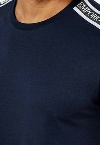 Mörkblå t-shirt med rundad halsringning, prydd med ett vitt och svart märke på axeln. Tillverkad av mjuk, lätt bomull.