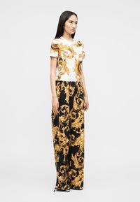 Γυναίκα που φοράει ταιριαστό λευκό και μαύρο τοπ και παντελόνι με φαρδιά πόδια με περίτεχνα χρυσά μπαρόκ σχέδια, στέκεται μπροστά σε ένα απλό φόντο.