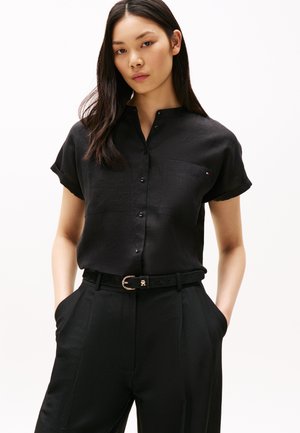 Chemise noire à boutons avec col mandarin et manches courtes, comportant deux poches avant et une texture subtile. Portée avec un pantalon noir et une ceinture.