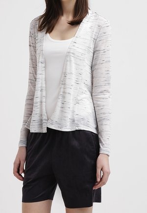 Kobieta w białym topie na ramiączkach, jasnoszarym kardiganie z otwartym przodem z czarnymi paskami oraz czarnych krótkich spodenkach do kolan, na tle jednolitego tła.