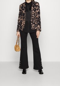 Blazer noir avec motif abstrait rose, top noir, pantalon noir évasé avec accents à pression et un sac à main beige froncé. Bottines noires.