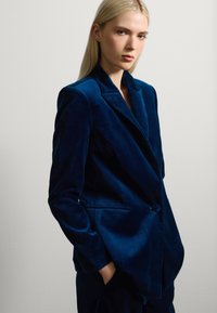Blazer de terciopelo azul oscuro con solapa con muesca, cierre de un solo botón y bolsillos laterales. Textura suave y ajuste entallado, que irradia un diseño clásico.