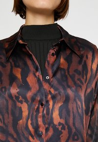 Brązowa i czarna koszula w wzór z satynowym wykończeniem, z kołnierzykiem w kształcie stalówki i zapięciem na guziki. Pod spodem czarna dzianinowa bluzka z wysokim kołnierzem.