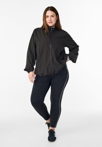 Schwarze Jacke mit Reißverschluss, langen Ärmeln und elastischen Bündchen, kombiniert mit schwarzen Leggings mit weißen Seitenstreifen und schwarzen Schnürschuhen.