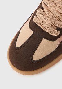 Κοντινό πλάνο μύτης καφέ και μπεζ σουέντ αθλητικού παπουτσιού με υφασμένες μπεζ κορδόνια και ελαφριά σόλα γκαμ σε λευκό φόντο.