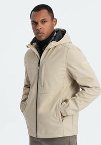 Beige Kapuzenjacke mit Reißverschluss, seitlichen Taschen, elastischen Bündchen und glatter Textur; darunter ist ein karierter Hemd zu sehen.
