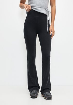 Femme portant un pantalon noir évasé à taille haute et un débardeur gris, soulevant légèrement sa chemise, debout devant un fond blanc uni.