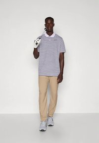 Mies, który ma na sobie w paski koszulkę polo, beżowe spodnie, białą rękawiczkę, trzyma kij golfowy na ramieniu i stoi na tle jasnego, gładkiego tła.