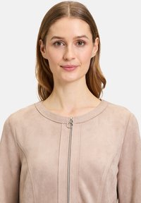 Ljust beige mockaskinnjacka med rund halsringning, dragkedja framtill med en silverring att dra i, samt subtila sömnadsdetaljer längs boddelen.