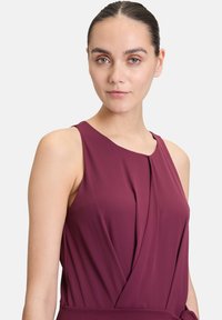 Robe sans manches bordeaux avec un buste drapé et un tissu doux. Elle présente un col rond et de légères pinces sur le devant.
