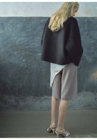 Zwarte cropped wollen trui, lichtgrijze gelaagde top, beige op maat gemaakte shorts en beige strap-schoenen, tegen een gestructureerde grijze achtergrond.