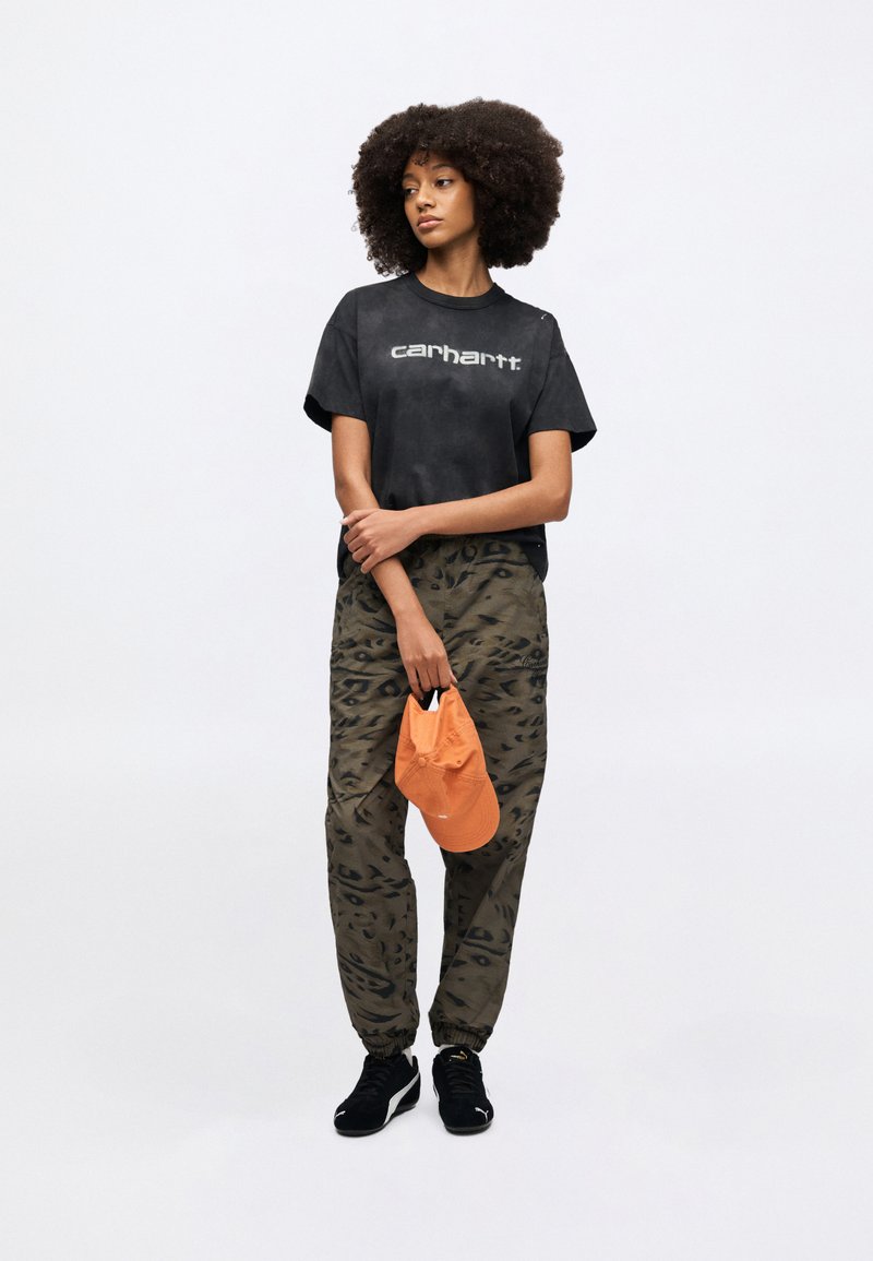 Sort bomuld T-shirt med "Carhartt"-logo, kombineret med olivengrønne cargo-bukser med mønster. Orange kasket holdt i hånden; sorte sneakers.