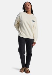 Φούτερ εκρού με στρογγυλή λαιμόκοψη και μανίκια με μανσέτες, σε συνδυασμό με σκούρο τζιν και καφέ σανδάλια με λουράκια. Διαθέτει μικρό λογότυπο.