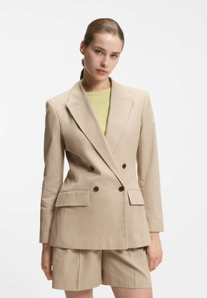 Kvinde med tilbagelagt hår iført en beige dobbeltradet blazer med nålestriber og matchende shorts over en lysegrøn top.