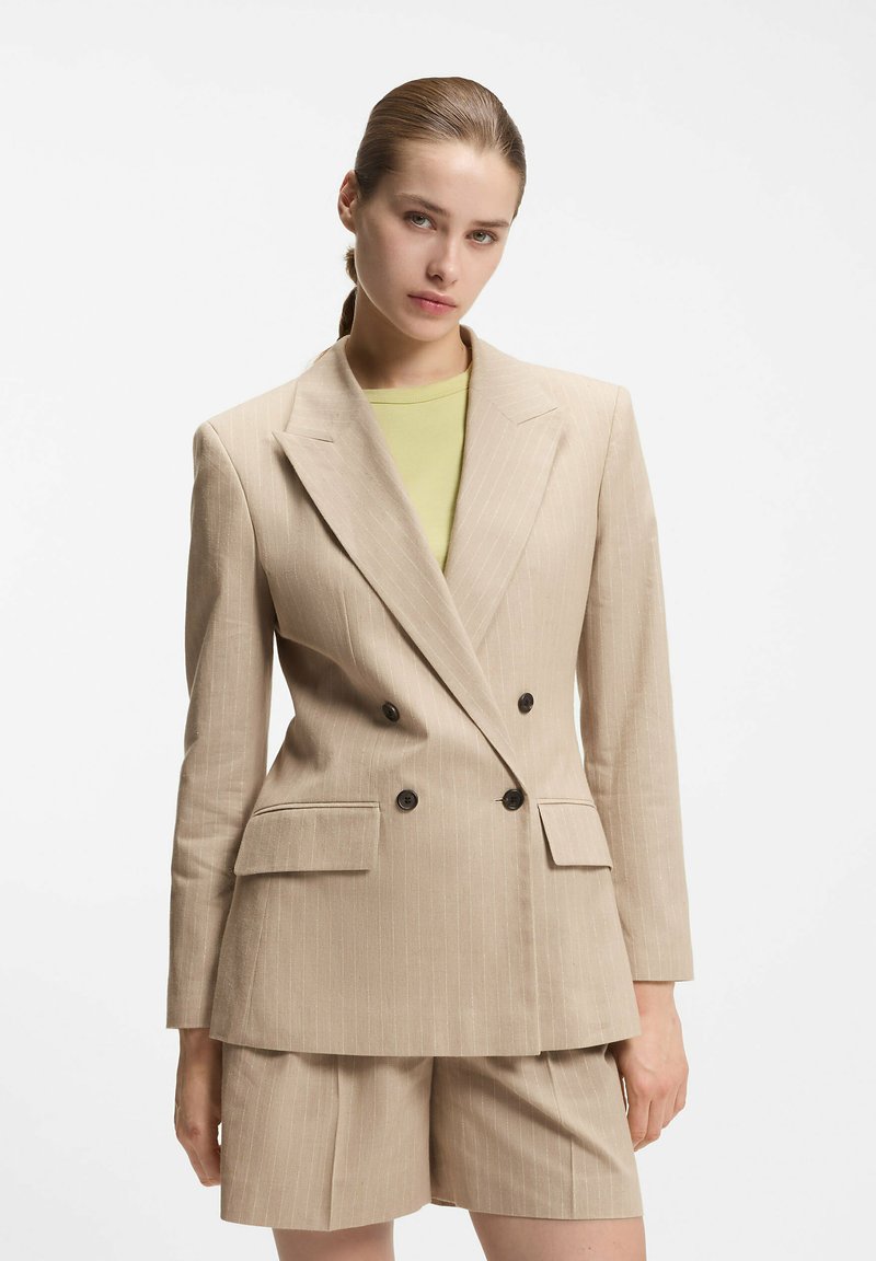 Kobieta z zaczesanymi do tyłu włosami, ubrana w beżową marynarkę dwurzędową w prążki i pasujące krótkie spodenki, na jasnooliwkowym topie.