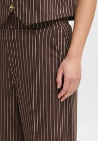 Haut en tissu brun à motifs avec des rayures blanches, présentant une poche poitrine et des détails de boutons, associé à un pantalon rayé brun. Main visible.