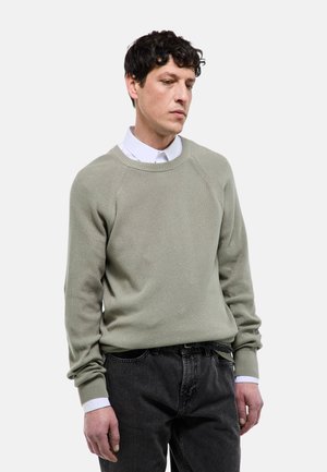 Homme aux cheveux courts et bouclés portant un pull en tricot gris clair sur une chemise blanche à col et un jean noir, debout devant un fond blanc.