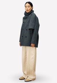 Manteau croisé bleu et gris à carreaux avec col haut, associé à un pantalon large crème et des chaussures beiges.