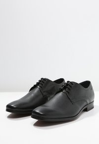 Une paire de chaussures en cuir noir pour hommes, formelles, avec un design perforé et des lacets, placées côte à côte sur une surface blanche.