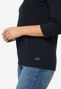Himmelblaues Langarmshirt aus weichem Stoff mit Rundhalsausschnitt und kleinem gesticktem Logo auf der unteren rechten Seite. Kombiniert mit hellblauen Jeans.