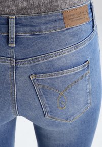 Jean en denim bleu clair à taille haute, avec une étiquette en cuir beige et un motif de couture dorée décoratif sur la poche arrière.