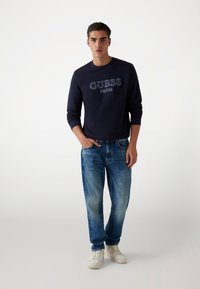 Felpa blu navy con logo "GUESS" in grigio chiaro; abbinata a jeans denim chiari; modello che indossa sneakers bianche.