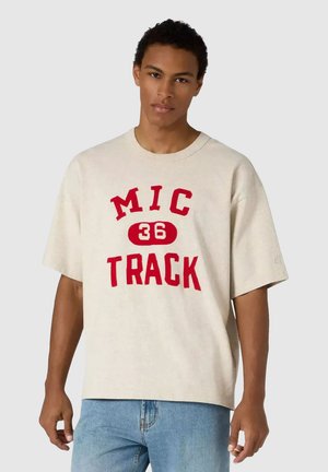 Jonge man draagt een beige oversized T-shirt met rode "MIC 36 TRACK" tekst en lichtblauwe jeans, staand tegen een effen achtergrond.