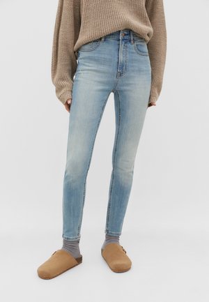 Personne portant un jean skinny taille haute bleu clair, un pull en maille beige, des chaussettes grises et des chaussures marron sans lacets sur un fond uni.