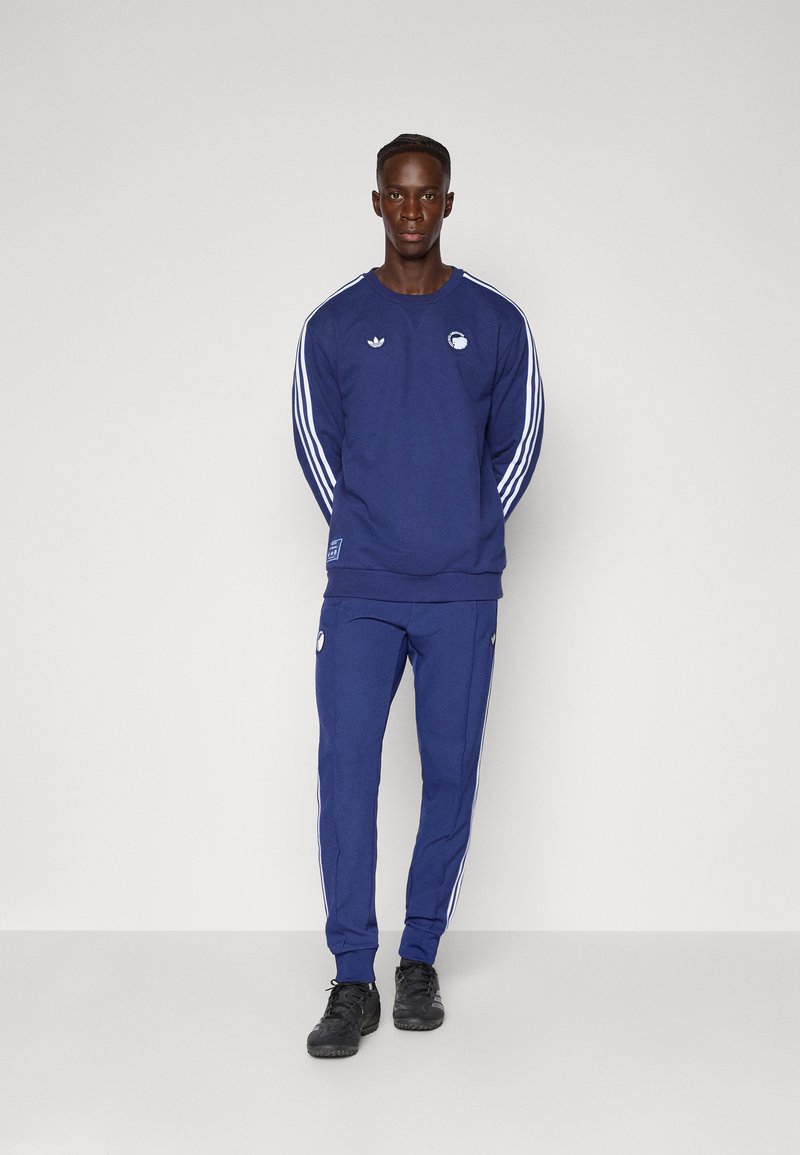 Chándal deportivo de color azul marino con franjas blancas, con cuello redondo y detalles de logo, combinado con zapatillas deportivas negras.